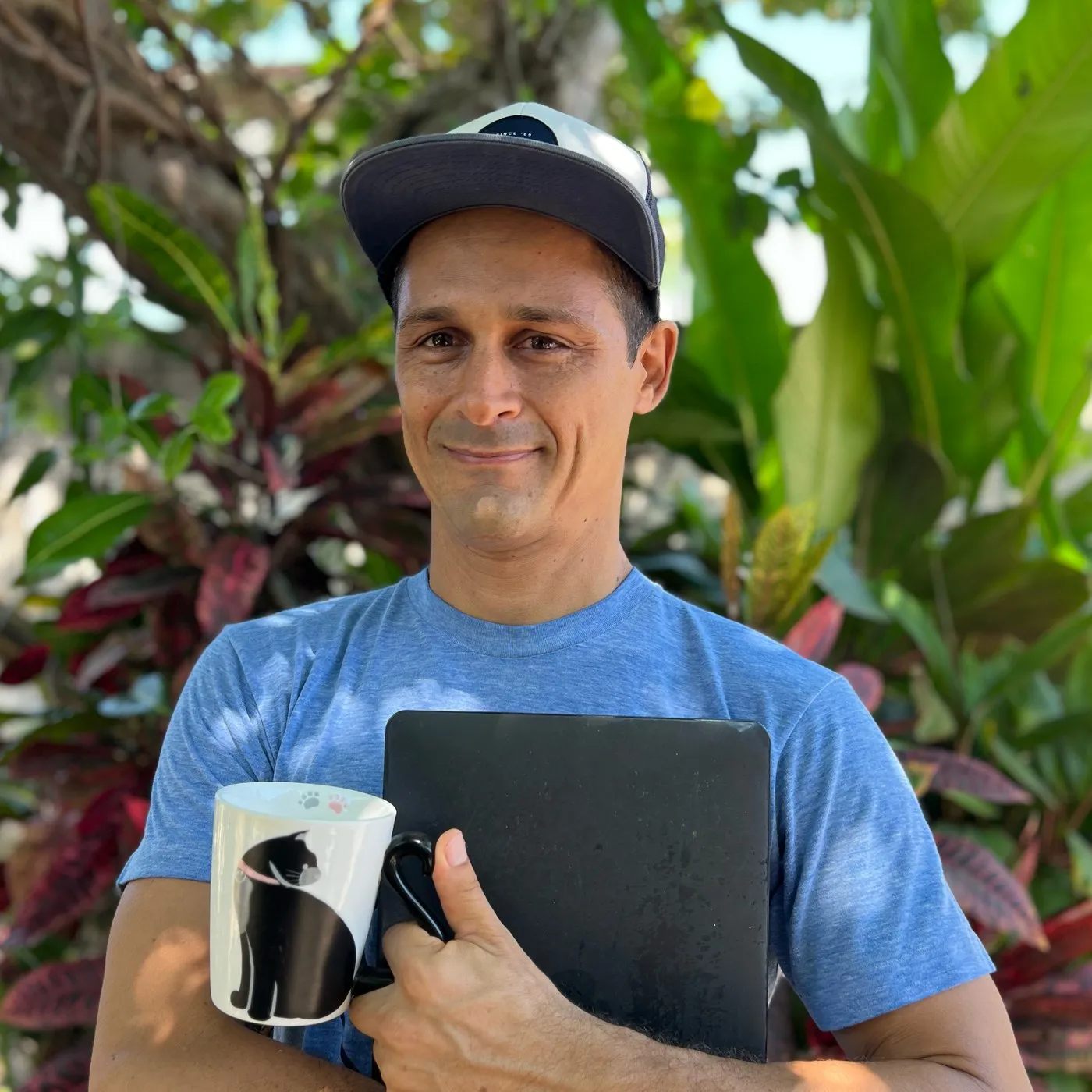 Gibran Garcia outdoors among tropical plants, holding a laptop and a coffee mug